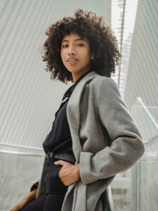 Modetrend Trenchcoat Frau mit Korkenzieherlocken trägt einen grauen Mantel zur schwarzen Jeans und Oberteil
