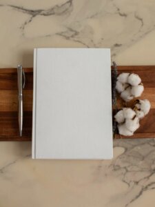 Weißes Notizbuch auf einem Holzbrett, daneben Stift und Baumwolle als Deko