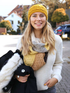 Warmes Outfit Mädchen trägt Stirnband, Pullover, Schal und eine Winterjacke auf dem Arm