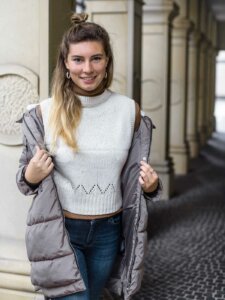 Winter Outfit Mantel Junge Frau trägt einen grauen Mantel, einen weißen Pullunder, ein braunes Rollkragenshirt und eine blaue Jeans