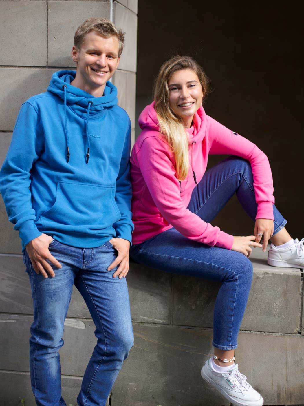 Frau im pinken Hoodie sitzt auf einer niedrigen Mauer, junger Mann im blauen Hoodie steht neben ihr.