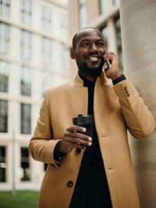 Mann trägt schwarzes Oberteil und beigefarbenen Mantel und hält Telefon und Kaffee in der Hand