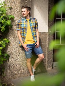 Junger Mann in Jeansbermuda, gelbem Shirt und kariertem Hemd lehnt an einer Hausfassade