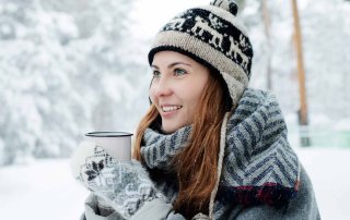 Winterlich angezogene Frau steht mit einer Tasse in den Händen in einer verschneiten Baumlandschaft