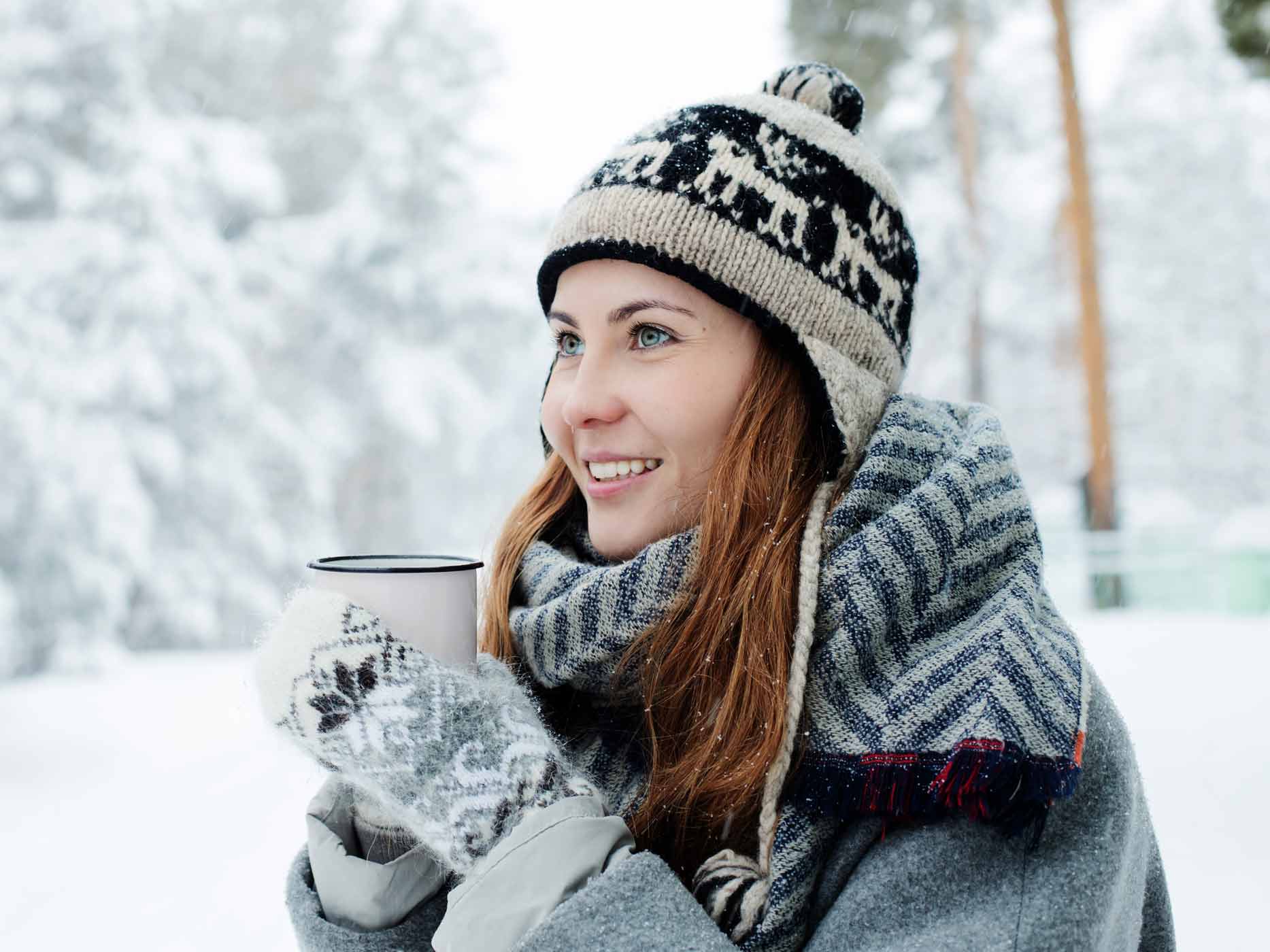 Winterlich angezogene Frau steht mit einer Tasse in den Händen in einer verschneiten Baumlandschaft