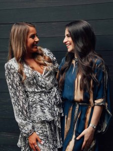 Weihnachtsoutfit Damen Kleid Zwei lachende Frauen in Kleidern stehen einander zugewandt vor einer Wand