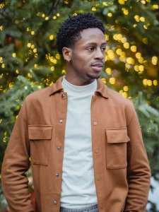 Weihnachtsoutfit Herren Cordhemd Dunkelhäutiger Mann vor Tannenbaum stehend mit weißem Pullver und brauner Jacke