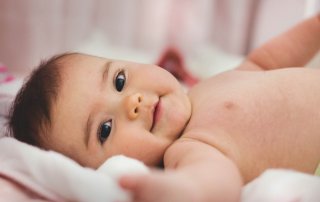 Ein Baby mit nacktem Oberkörper liegt auf einer kuscheligen Decke und lächelt in die Kamera