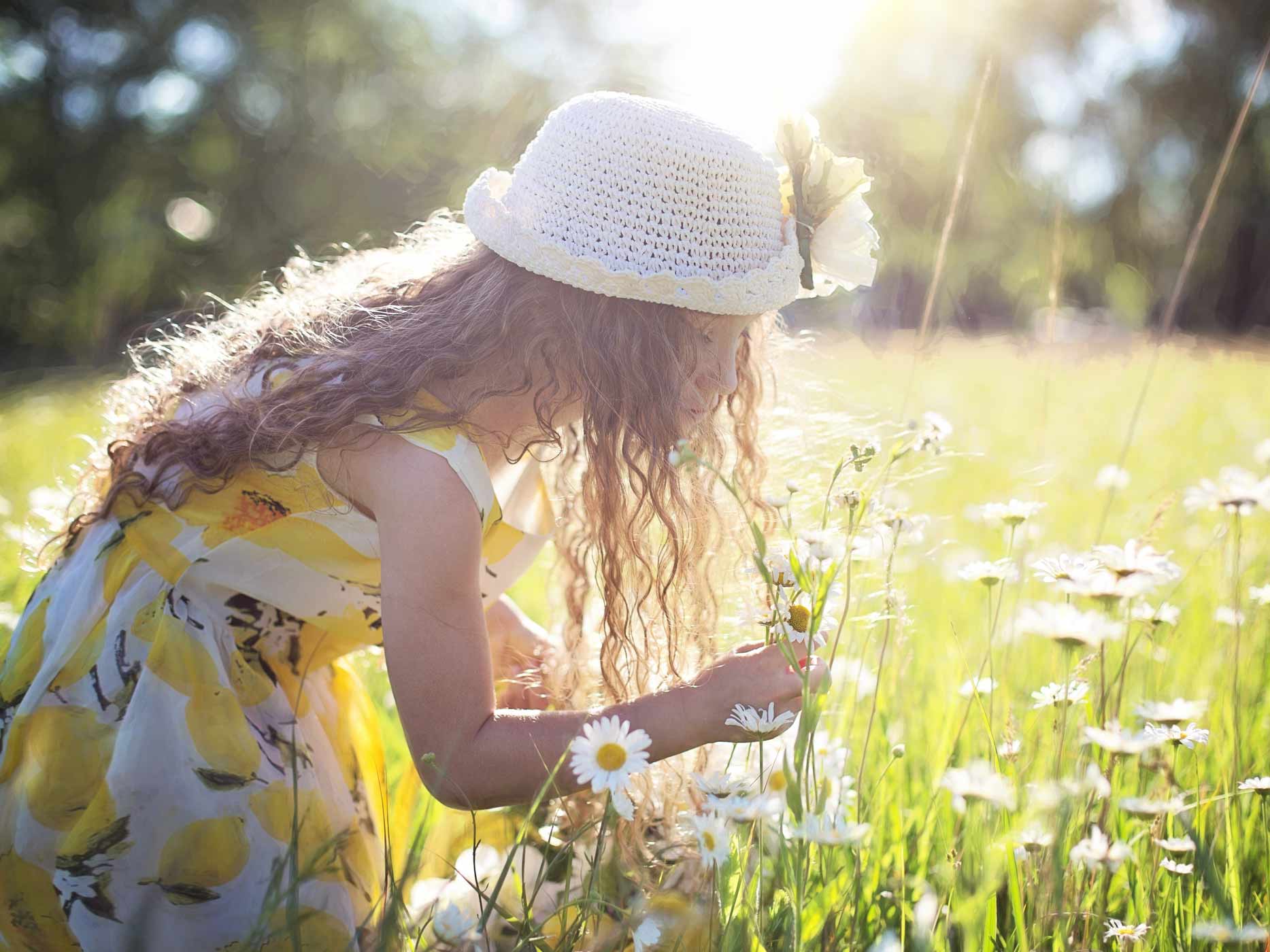 Mädchen im Sommerkleid und mit Hut pflückt auf einer sonnigen Wiese Blumen