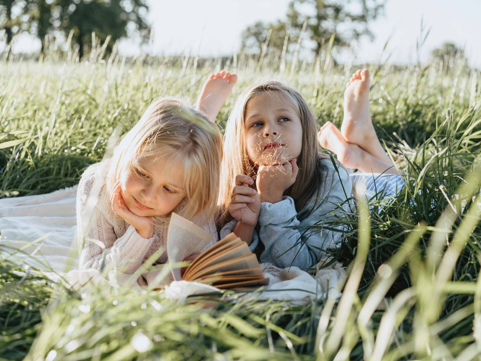 Zwei Mädchen liegen im Gras einer sonnigen Wiese und lesen