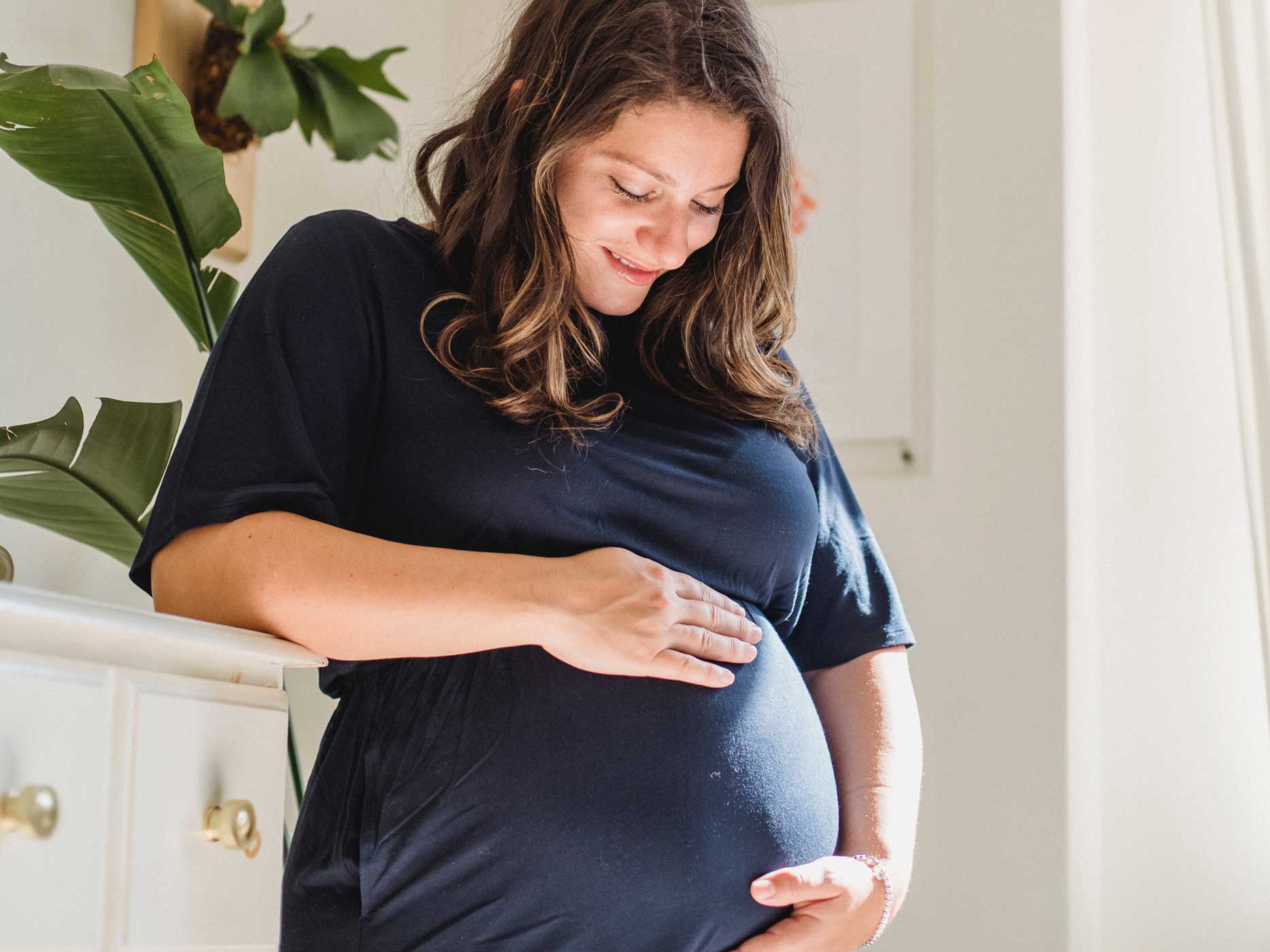 Eine werdende Mutter im dunkelblauen Kleid streichelt glücklich ihren Babybauch.