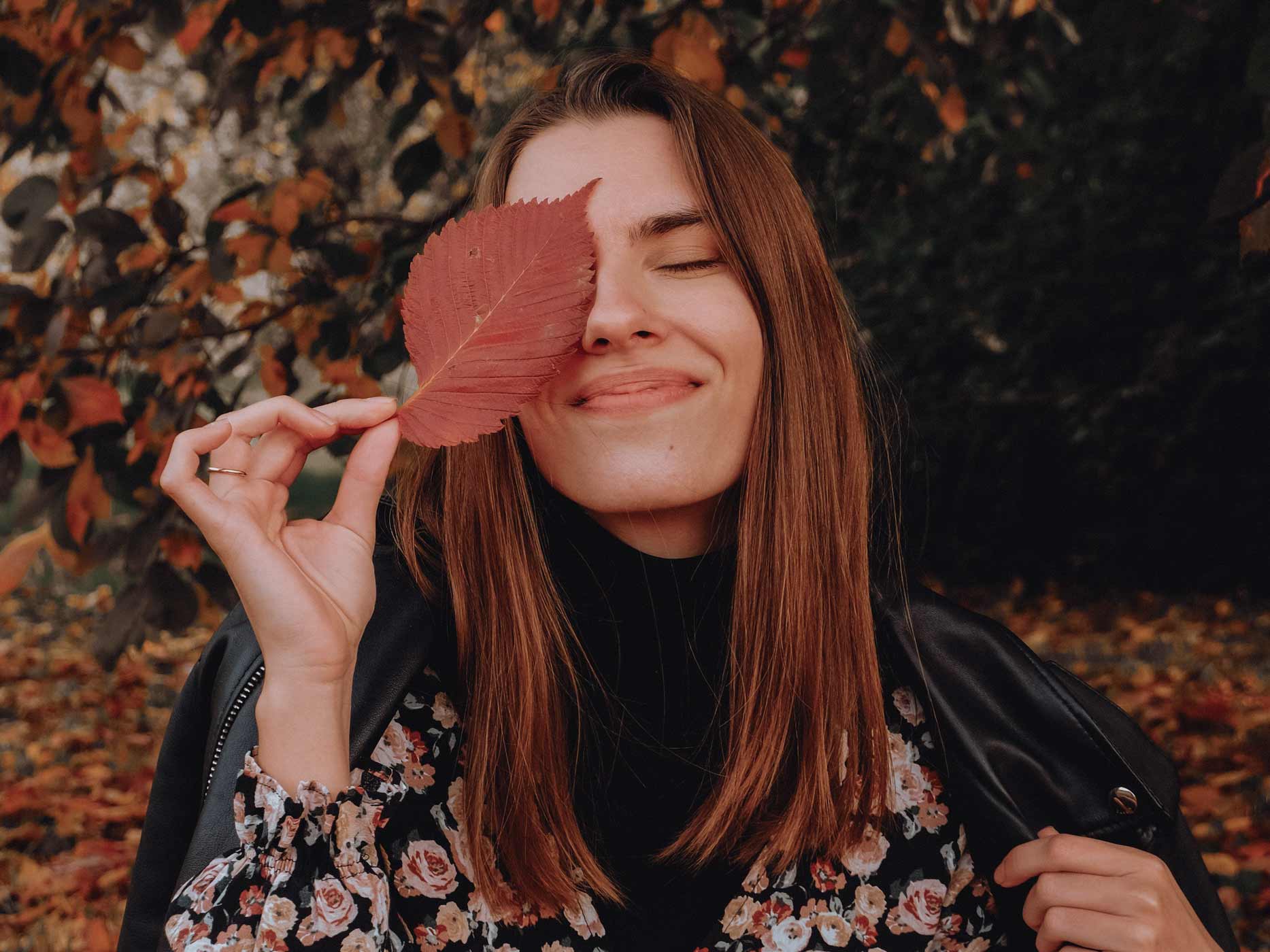 Frau vor herbstlich-verfärbtem Laub hält sich ein rotes Blatt vors Auge