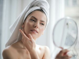 Frau trägt ein Handtuch auf dem Kopf und betrachtet ihr Gesicht in einem Kosmetikspiegel in ihrer Hand
