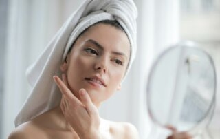 Frau trägt Handtuch auf dem Kopf und betrachtet ihr Gesicht in einem Kosmetikspiegel in ihrer Hand