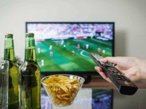 Fußball EM Tipps Fernbedienung wird auf einen Fernseher gerichtet, der Fußball zeigt. Bierflaschen und Chips stehen auf einem Tisch.