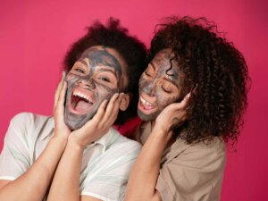 Zwei Frauen lächeln in die Kamera, sie tragen eine Gesichtsmaske und stehen vor einer pinken Wand.