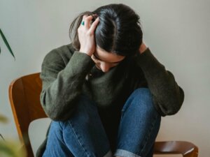 Eine Frau kauert sich auf einem Stuhl zusammen und hält den Kopf verzweifelt in beiden Händen.