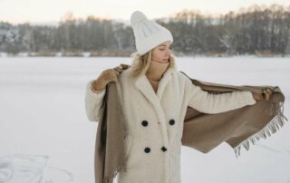 Eine Frau steht in einem weißen Teddymantel mit Mütze, Schal und Handschuhen vor einer Schneelandschaft.