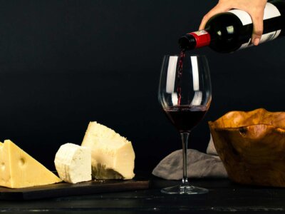 In ein Glas wird Rotwein eingeschenkt, drum herum liegen verschiedene Arten von Käse.
