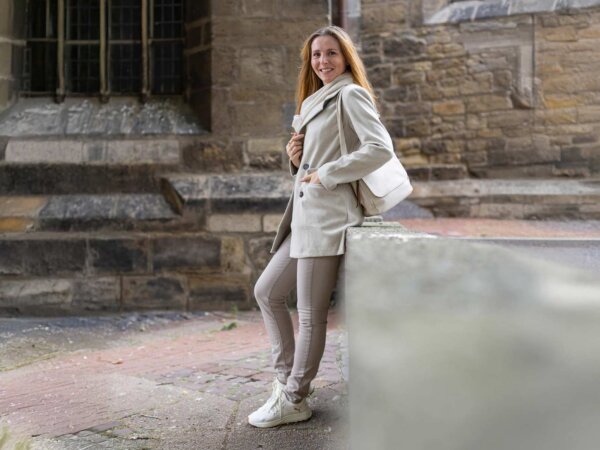 Eine Frau lehnt an einer Mauer und trägt ein beiges, monochromes Outfit. Im Fokus steht dabei ein Trenchcoat.