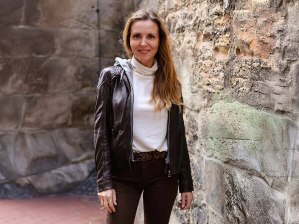 Eine Frau trägt eine braune Hose, ein weißes T-Shirt und eine braune Lederjacke.