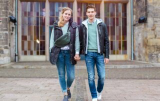 Eine Frau und ein Mann gehen händchenhaltend spazieren, sie tagen Schwarze Westen/Jacken und mintfarbene Oberteile dazu.