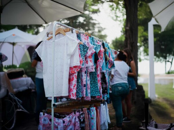 Auf einem Flohmarkt steht eine Kleiderstange, auf der mehrere bunte Damenoberteile mit Kleiderbügeln aufgehängt sind.