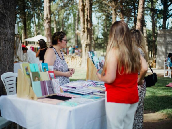 Eine Frau hat einen Tisch auf einem Flohmarkt, der draußen stattfindet. Vor ihrem Stand stehen zwei Frauen, die sich von der Frau etwas erklären lassen.