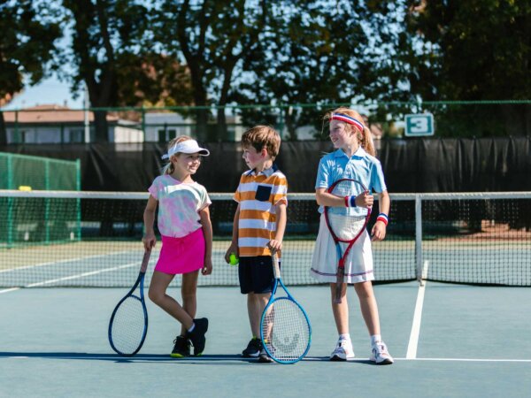 Ein Junge und zwei Mädchen stehen in Sportkleidung und mit Tennisschlägern auf einem Tennisfeld.