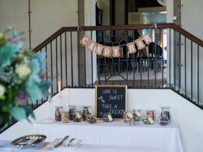 Ein Tisch ist mit einem weißen Tuch bedeckt, darauf sind verschiedene Gläser mit Süßigkeiten gefüllt. Darüber hängt eine Girlande mit der Aufschrift "Candy Bar". 