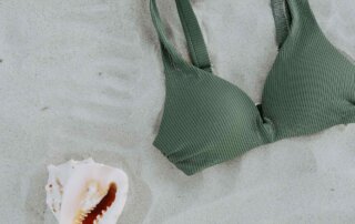 Ein grünes Bikinioberteil liegt auf Sand und daneben eine weiße Muschel.
