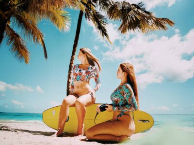 Zwei Frauen sitzen auf einem Surfbrett am Strand unter Palmen und tragen dabei bunte, sportliche Bikinis.