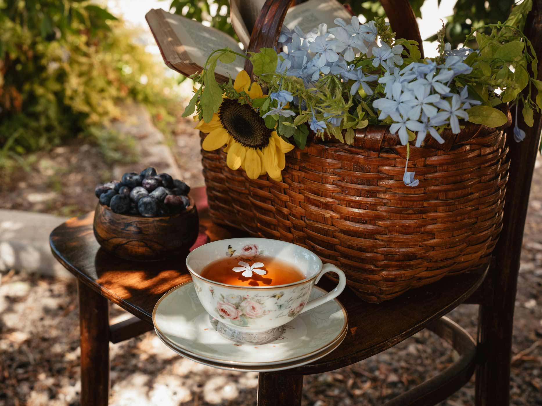 Auf einem Stuhl im Garten steht ein Korb mit Blumen, eine Schale mit Blaubeeren und eine Teetasse.