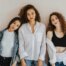Drei Frauen stehen vor einer weißen Wand, eine trägt eine schwarze Jeans, ein weißes T-Shirt und eine Jeansjacke, die Frau in der Mitte eine hellblaue Jeans und eine langärmelige weiße Bluse und die Frau rechts eine blaue Jeans, ein schwarzes BH-Top und ein weißes Hemd.