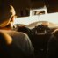 Blick vom Rücksitz eines Autos aus auf den entspannten Fahrer mit Cap, der gegen die Sonne fährt.
