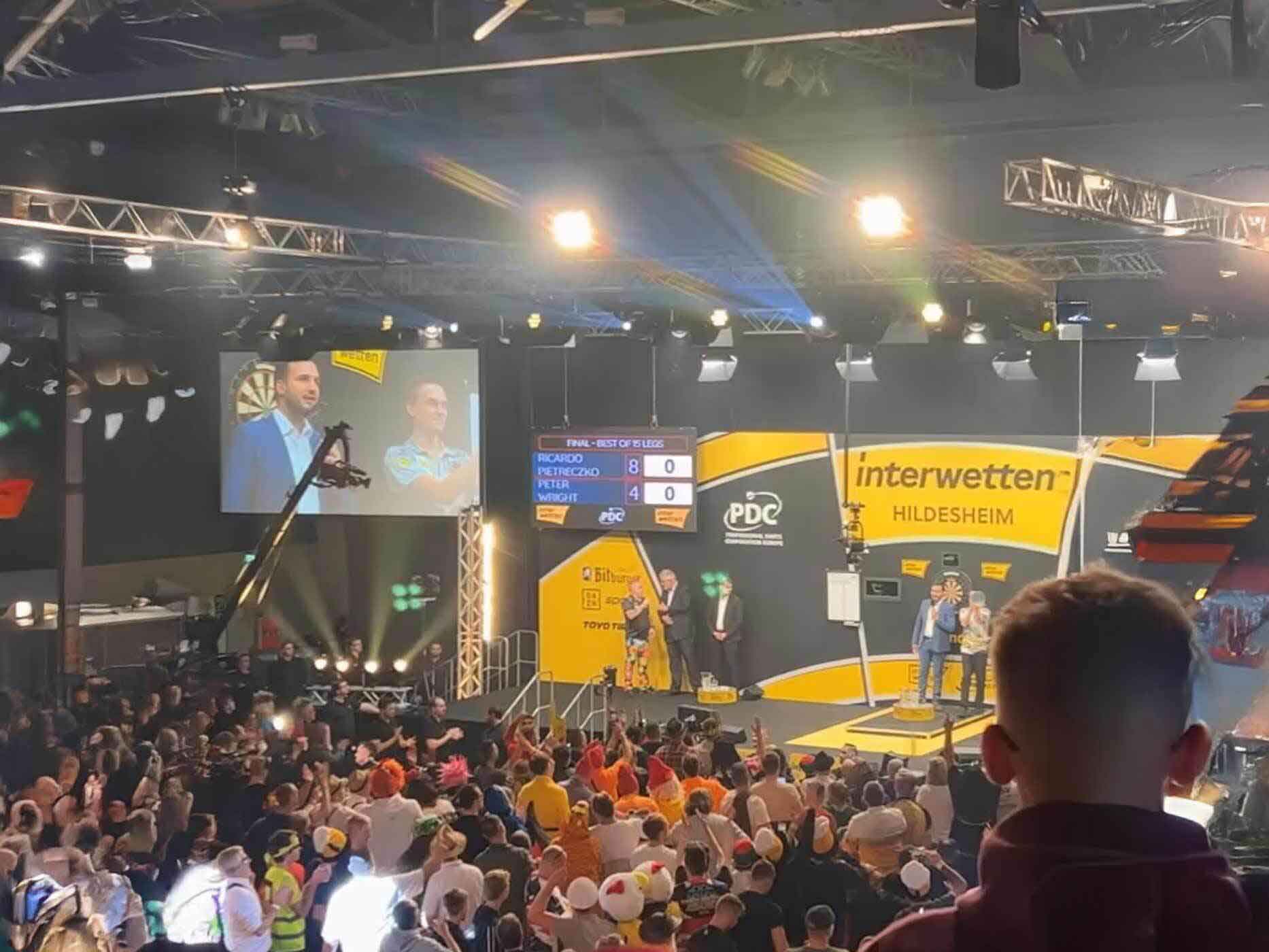 Darts Championship Hildesheim Vogelperspektiveaufnahme der Halle 39, die Fans stehen und Jubeln, auf der Bühne der Moderator und ein Darts Spieler.