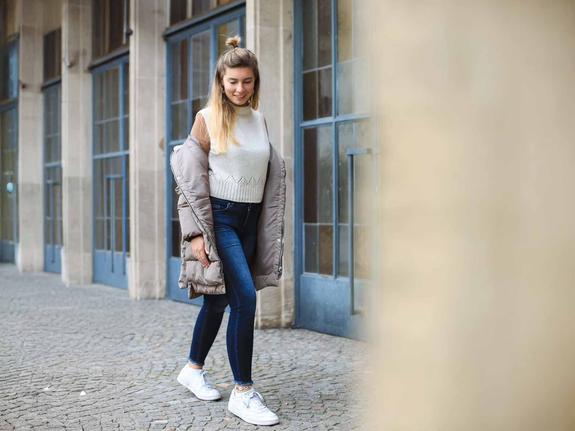 Look aufwerten Details Eine Frau trägt eine dunkelblaue Jeans, ein braunes Langarmshirt und darüber einen beigen Pullunder. Darüber trägt sie eine offene grau-beige Jacke und weiße Sneaker.