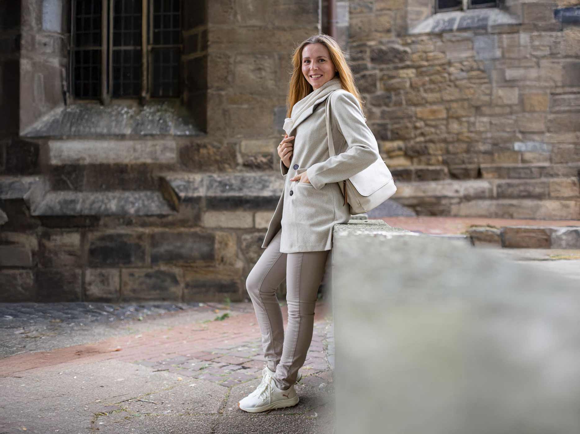 Look aufwerten monochrom Eine Frau lehnt an einer Mauer, sie trägt eine Jeans, einen Mantel, einen Schal und eine Handtasche alles im gleichen beige-grauen Farbton. Dazu weiße Sneaker.